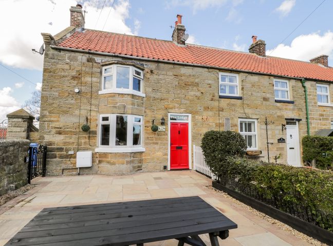 Stone house with a red door and bay window a black picnic table and bushes at The Lodge in Stainsacre near Whitby