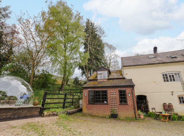 An outdoor area with a small brick building and a white building next to it with chairs and tables surrounded by trees at The Wheelhouse at Dudgeley Mill in All Stretton