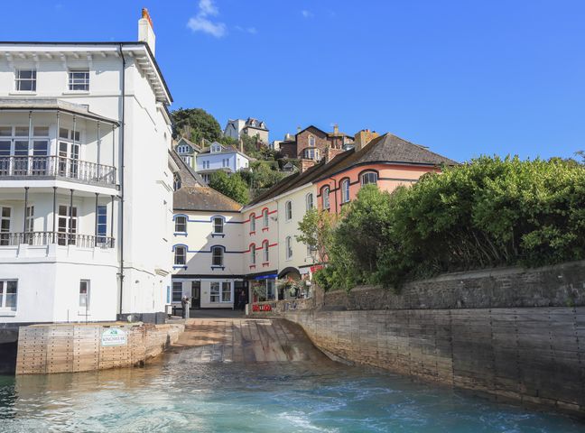 A waterfront scene with multi-story buildings near a boat ramp and greenery at Ferrystone in Kingswear near Dartmouth