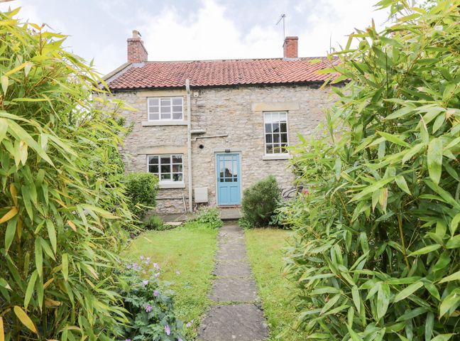A stone house with a blue door and two chimneys seen from a garden with a stone path and green plants at Cliff House in Helmsley