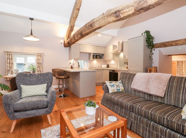 An open plan living room and kitchen with wooden beams a striped sofa a grey armchair a glass coffee table and bar stools at Low Ashmead Cottage in Hawkshead