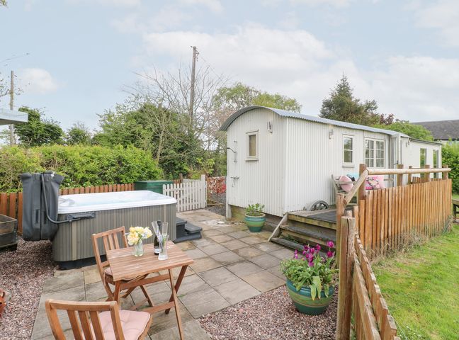 An outdoor patio with a wooden table and chairs a hot tub and a small white corrugated metal cabin at Shepherds Hut in Colton near Rugeley Staffordshire