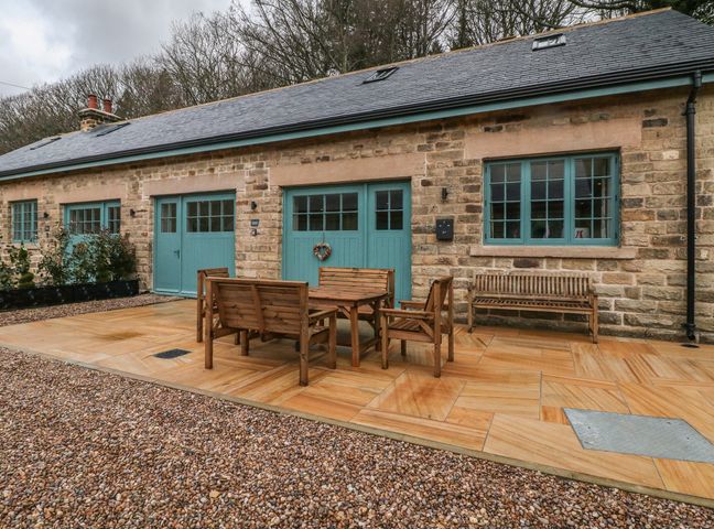 An outdoor patio with wooden table chairs and benches in front of a stone building with teal doors and windows at Chestnut Cottage in Stocksbridge