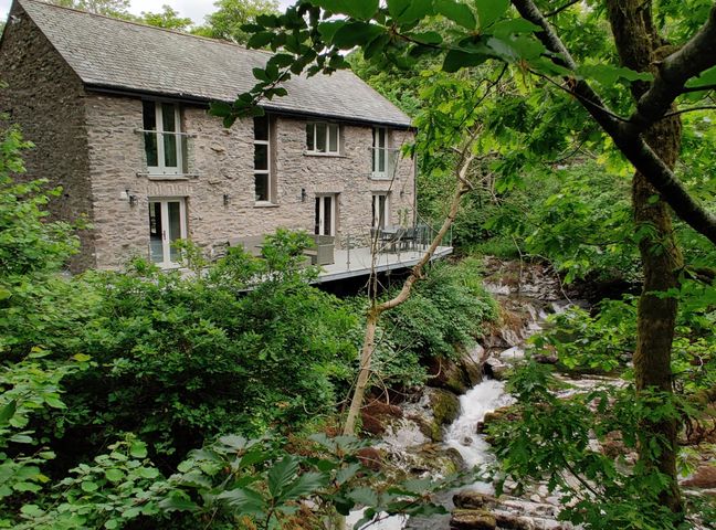 A stone house with multiple windows and doors next to a stream surrounded by trees at The Old Water Mill in Kendal