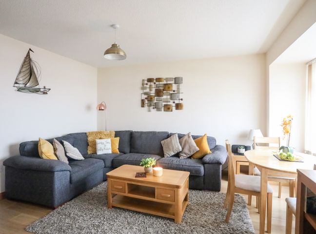 A living room with a dark sectional sofa and wooden coffee and dining tables at Sandy Toes in Rhosneigr
