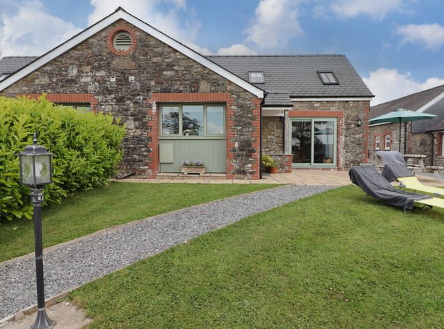 A stone cottage with a gravel path leading to the door and lounge chairs on the lawn at Barcud Cottage in Golden Grove near Llandeilo