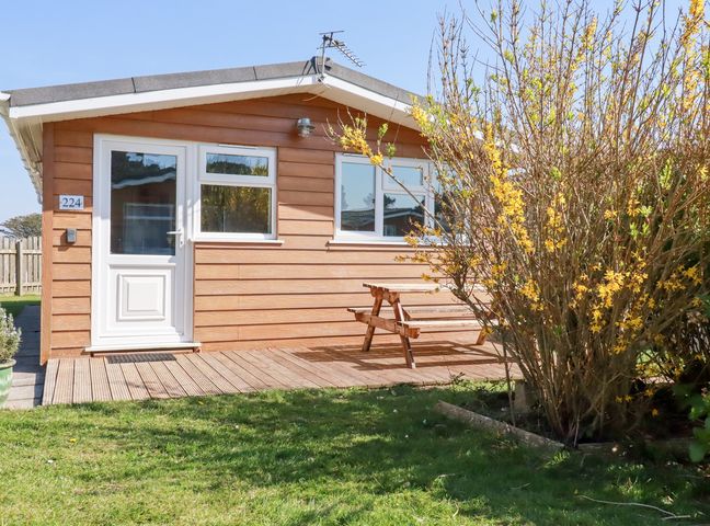 A wooden cabin with white door and windows a picnic table on a deck and a flowering bush in the yard at 224 Atlantic Bays in St Merryn
