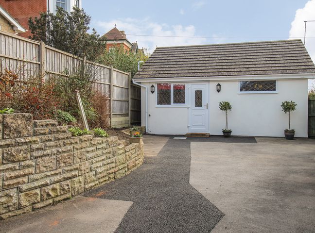 A small white building with a shingled roof and two small trees in pots on either side of the door at Roundham Cottage in West Bay