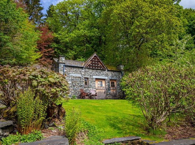 A stone cottage with wooden doors and chairs on a lawn surrounded by green trees at Dovecot Cottage in Grasmere
