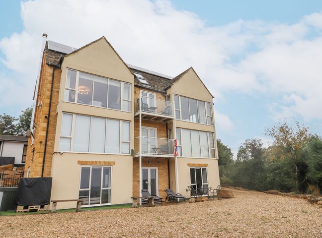 A multi-story building with large windows and balconies with outdoor chairs on a gravel area surrounded by trees at Cotswold View in Bourton-On-The-Water