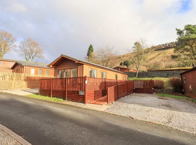 A wooden lodge with a red wooden ramp and railings next to a gravel parking area with trees and hills in the background at Lakeland View Lodge Beckside 2 Limefitt Park near Windermere
