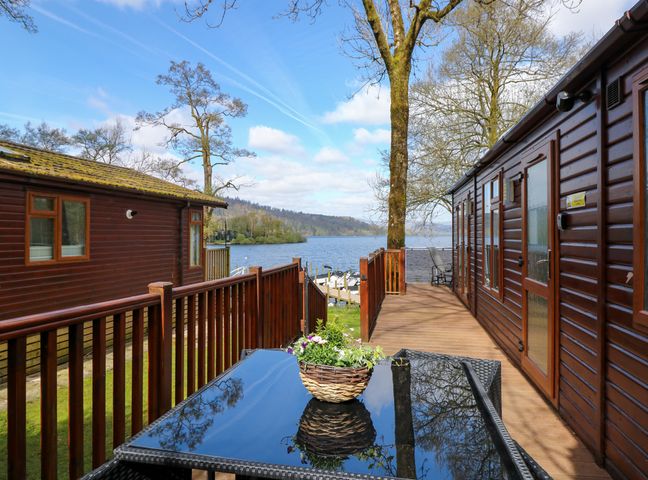 A deck with wooden railings and a table with a flower basket between two wooden lodges by a lake at Shoreside Lodge L8 Fallbarrow Park near Bowness on Windermere