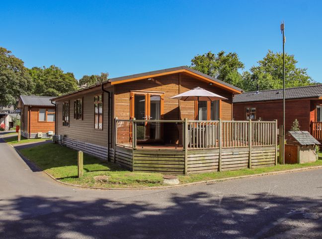 A wooden cabin with a fenced deck and umbrella surrounded by trees and other cabins at Daffodils Lodge Oakside 7 Fallbarrow Park in Bowness on Windermere