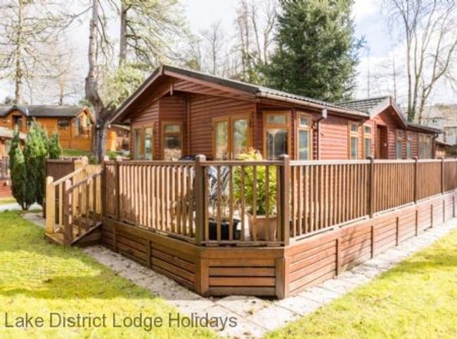 A wooden lodge with a fenced deck surrounded by grass and trees at Broad Larch Lodge Glade 16 Fallbarrow Park near Bowness on Windermere