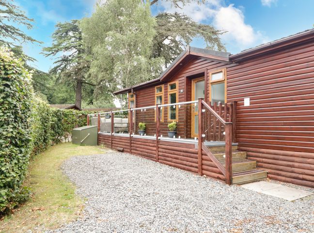 A wooden cabin with a glass railing and potted plants outside along a gravel pathway at Beech Grove Lodge in Glade 17 Fallbarrow Park near Bowness on Windermere