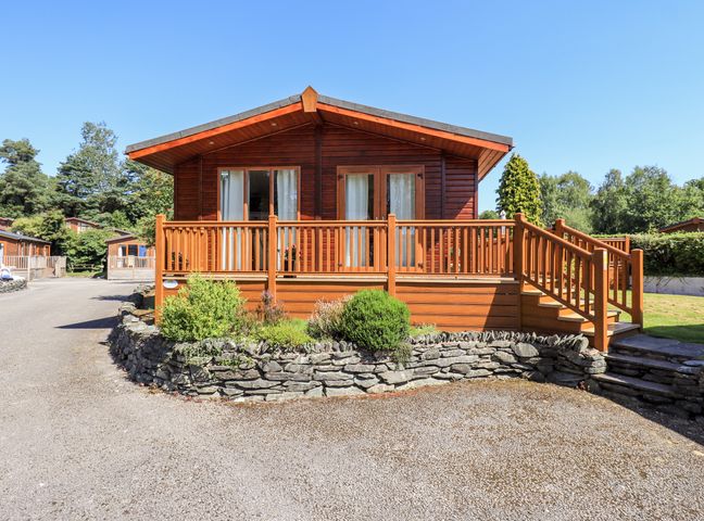 A wooden cabin with a deck and stairs surrounded by a stone wall and greenery at Robins Nest in Bowness 71 White Cross Bay near Windermere