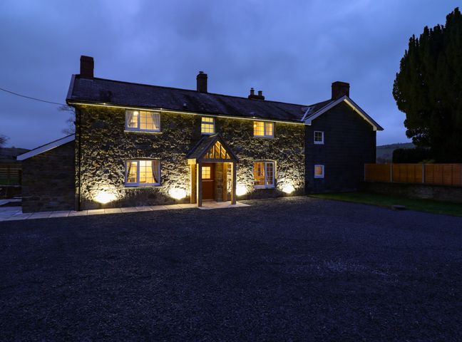 A stone cottage with multiple windows lit at night and a gravel driveway at Bodaioch Cottage in Trefeglwys near Caersws