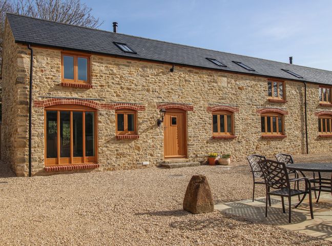 Exterior view of a stone house with multiple wooden windows and a wooden door next to outdoor dining furniture at The Granary Burton Bradstock