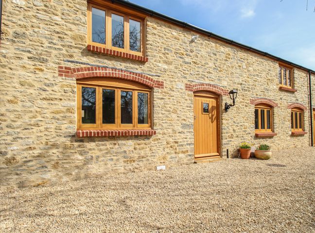 An exterior view of a stone house with wooden windows and door on a gravel ground at The Stables Burton Bradstock