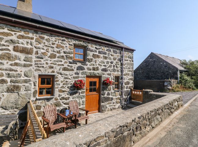 Exterior of a stone cottage with wooden door and windows two wooden chairs with a small table outside and flower baskets on the wall at Bwthyn Siop Pencaerau in Rhiw near Aberdaron