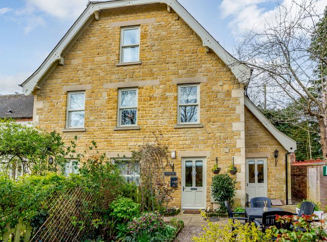 The front view of a two story stone house with multiple windows and doors and a garden with outdoor chairs and table at Jasmine Cottage Mickleton Gloucestershire