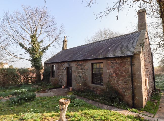 A stone cottage with two windows and a door surrounded by a garden and trees at Rose Cottage Akeld near Wooler