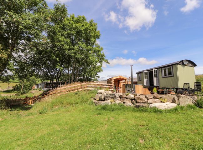 A small green cabin with open doors on a stone patio surrounded by chairs and a wooden fence near trees on a grassy hillside at The Moorhen Retreat in Nantmawr near Trefonen