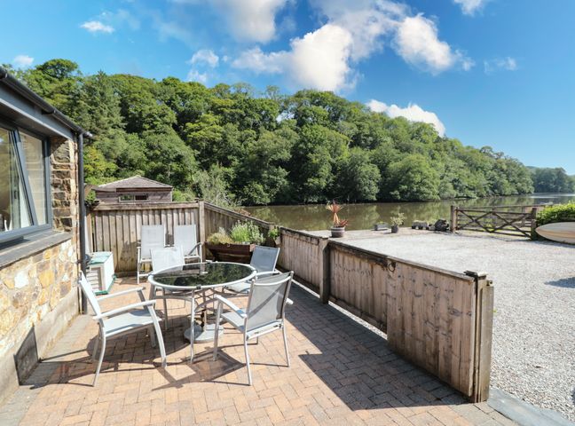 An outdoor patio area with a round glass table and six chairs next to a stone house by a river with trees in the background at Mallard Cottage in Totnes