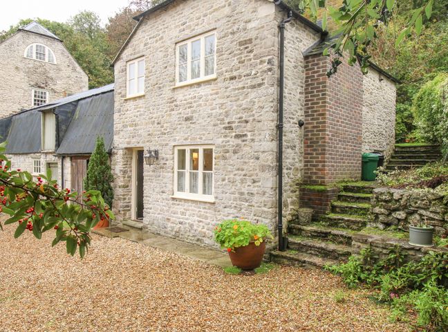 A stone cottage with two windows a door and potted plants on a gravel yard with stone steps and greenery at Mill Cottage in Upwey