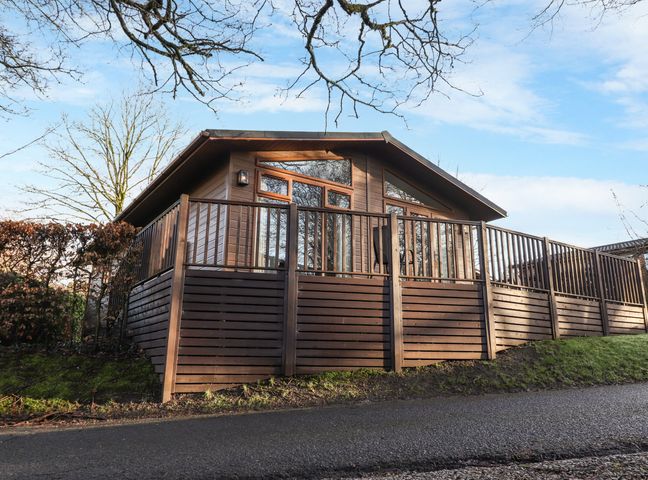 A wooden cabin with glass windows and a surrounding wooden fence on a grassy slope at Westbrook Lodge in R12 Fallbarrow Park near Bowness-On-Windermere