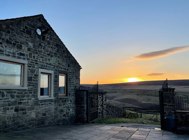 A stone building with three windows next to an open iron gate overlooking fields at sunset at The Salt Pot in Oakworth