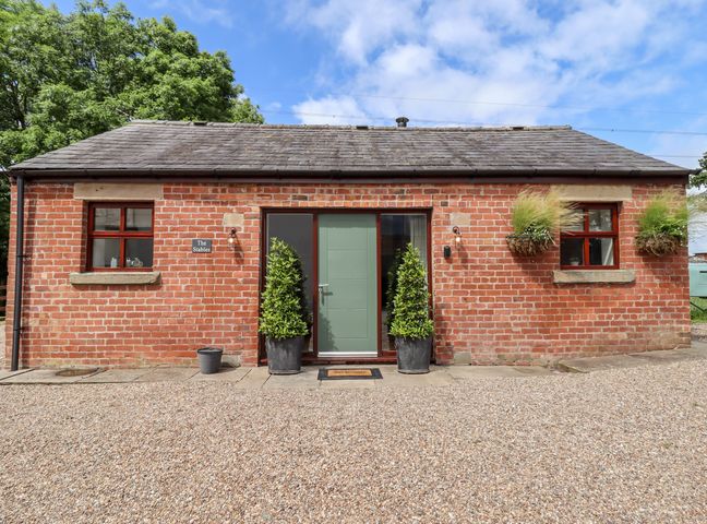 A single-story brick building with a green door two potted plants and two windows at The Stables Hall Green near Walmer Bridge