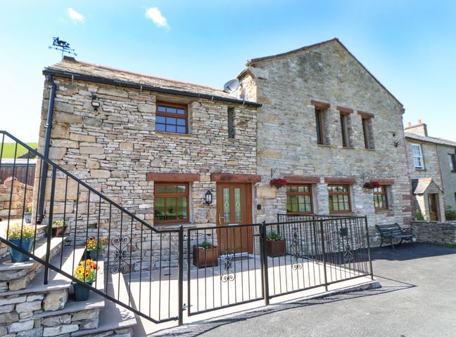 Exterior stone wall of a house with wooden framed windows and door with metal gate and stone steps with flowers at Birkett Barn in Kirkby Stephen