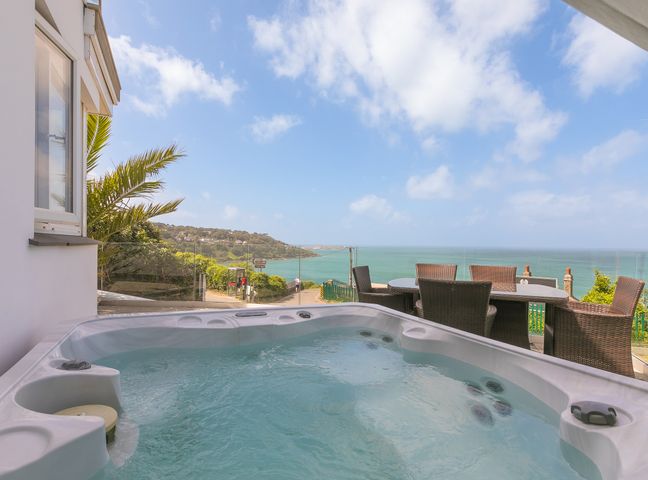 An outdoor hot tub and wicker dining chairs with a glass railing overlooking the sea at Starboard in Carbis Bay