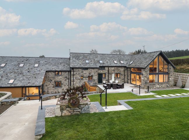 A stone house with slate roof and large windows overlooking a patio with outdoor furniture and green lawn at Merddwr in Cerrigydrudion