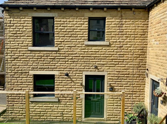 A stone house exterior with three windows and a green door surrounded by plants at Victoria House in Holmfirth