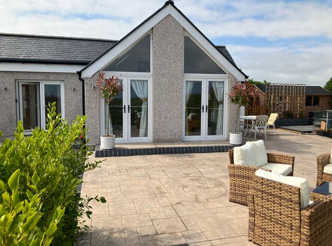 An outdoor patio area with wicker chairs with white cushions two potted trees a table with chairs and a house with triangular roof windows at Cherry Tree Cottage in Sandhead