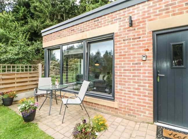 A patio with a glass table and two chairs next to a brick building with a dark door and large windows at Leadmill Piggery in Copley near Barnard Castle