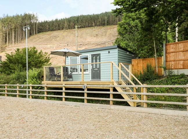 A small cabin with a deck and patio furniture surrounded by trees and a fenced area at Caban Glas in Llanafan near Pontrhydfendigaid
