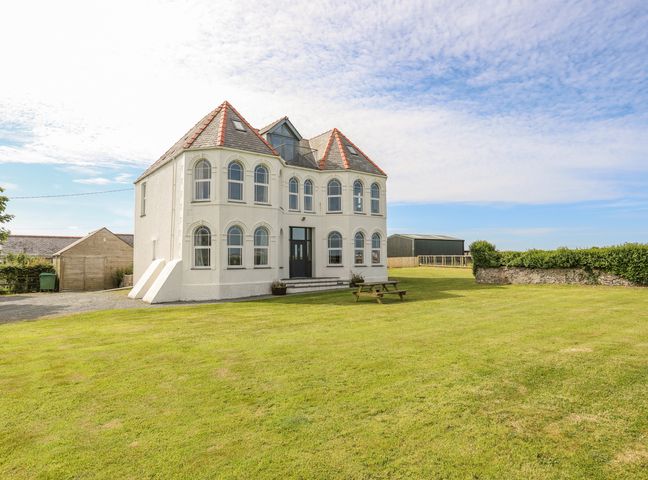 A two-story house with arched windows and a red slate roof on a lawn with a picnic table at Plas Newydd in Llanfaelog near Rhosneigr