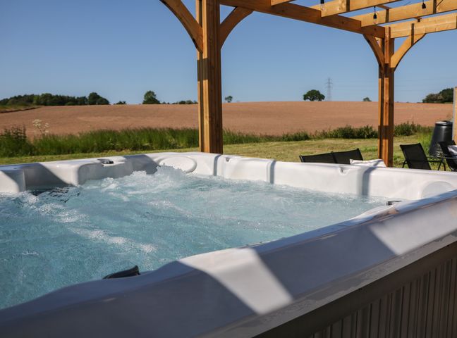 An outdoor hot tub with bubbling water under a wooden pergola with chairs and a field in the background at Langley Barn in Kirk Langley near Brailsford