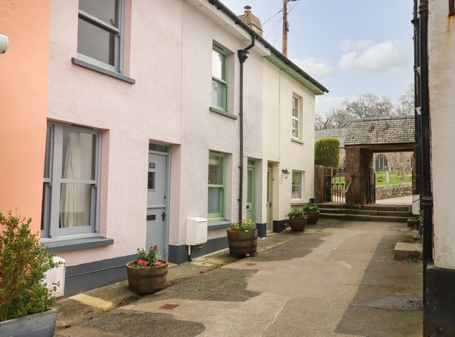 A narrow street with pastel-colored houses and flower pots leading to a stone archway at Church Cottage in Chagford
