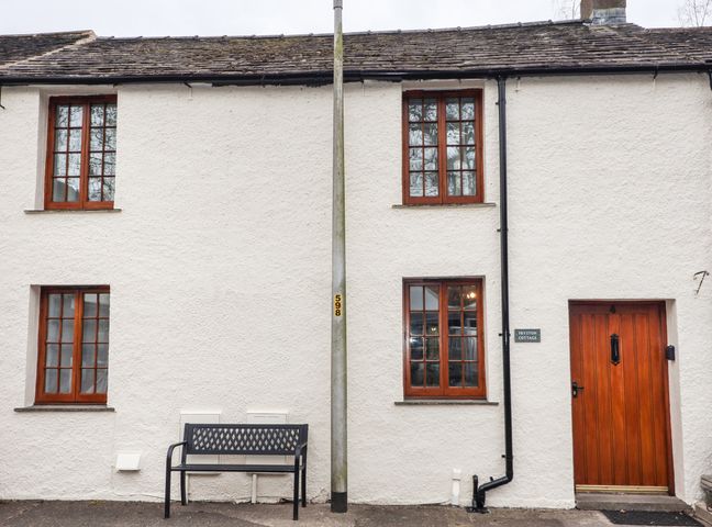A white cottage exterior with four wooden framed windows a wooden door a black bench and a central lamppost at Fryston Cottage in Keswick