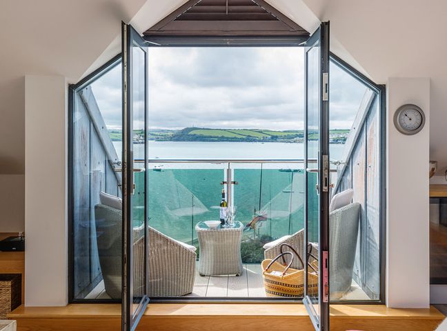 A balcony with wicker chairs a small table and a basket overlooking a body of water and distant hills at Upper Deck in Rock