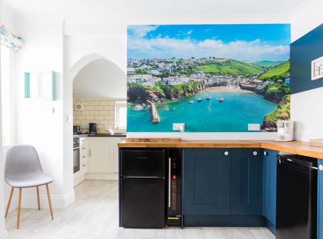 A kitchen with blue cabinets wooden countertops a black fridge a grey chair and a wall mural of a coastal town at Number 28 in Port Isaac