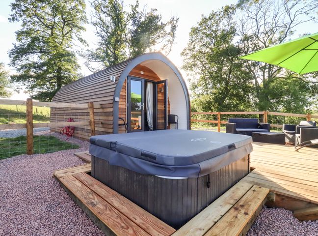 A wooden pod with double doors next to a hot tub on a wooden deck with seating and a green umbrella near trees at Goose Pod Near Clifton Upon Teme
