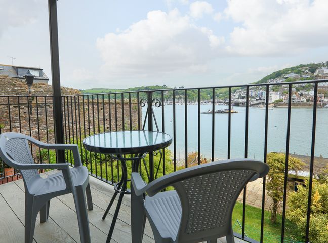 A balcony with a round glass table and two plastic chairs overlooking a river and houses on a hillside at Ferry View Dartmouth