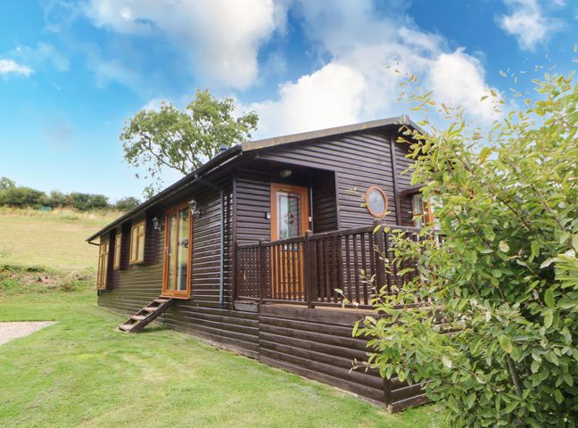 An exterior view of a dark wooden cabin with a small porch and wooden railing surrounded by grass and bushes at Badgers Retreat in Tunstall near Hipswell