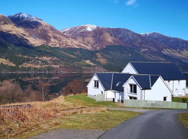 A white house with dark roof near a lake and mountains at Lochside 1 The Corries in Spean Bridge