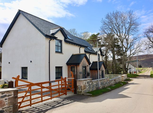 A white two-story house with stone walls and wooden gates on a street with trees at Kilmuir Cottage in Tomintoul